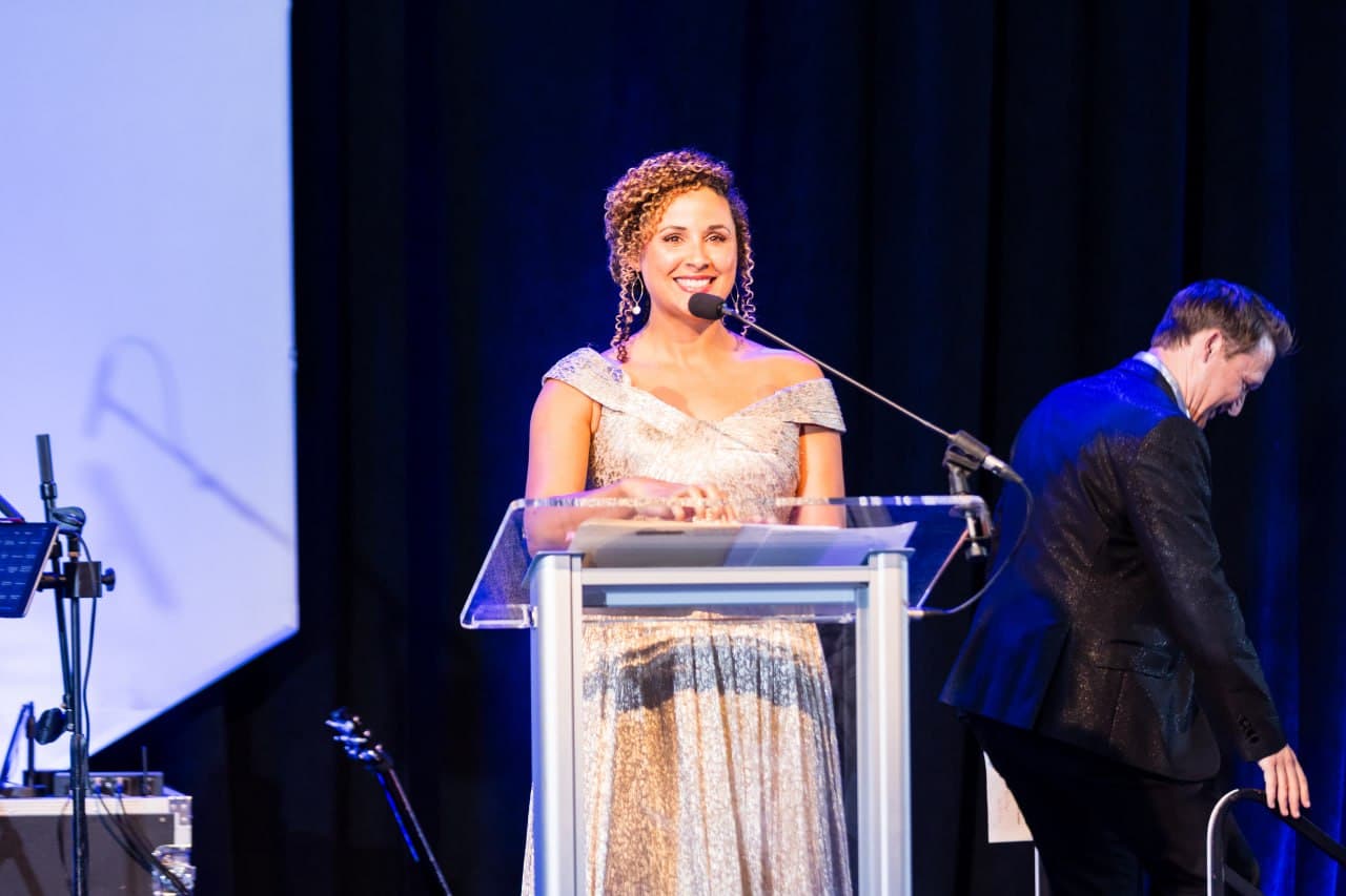 Christine Sylvain in gold sequin gown speaking at awards gala podium